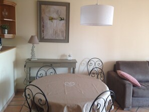 A dining area with a round table covered by a cloth, a metal chair, a sofa with a cushion, a lamp on a side table, and a painting on the wall.