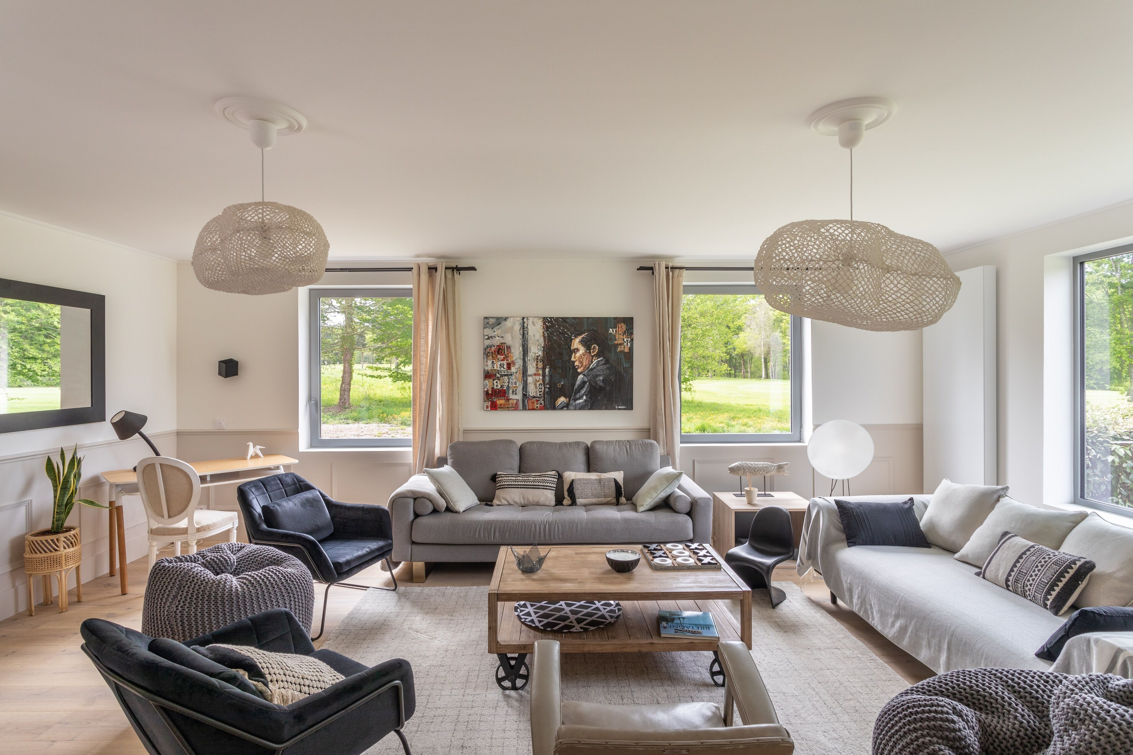 Een moderne woonkamer met een grijze bank, zwarte fauteuils, een houten salontafel en grote ramen met uitzicht op groen.