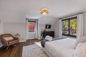 A bedroom with a large bed, a brown leather chair, a small side table, a TV on the wall, and a window with a view of trees.