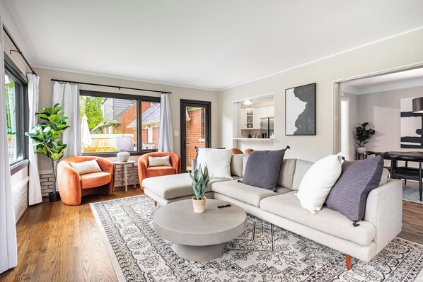 A modern living room with a large sectional sofa, a round coffee table, and two armchairs. There is a large window providing natural light and a view of the outside.