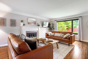 A living room with a fireplace, a brown leather sofa, a coffee table, and a large window with a view of greenery outside.