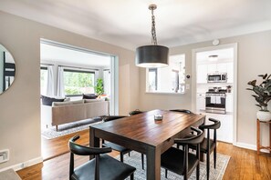A modern kitchen with a wooden dining table and chairs, a hanging light fixture, and a view of the outdoors through large windows.