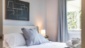 A neatly made bed with white and gray bedding, a wall art piece, a bedside lamp, and a window with curtains. - Spacious, Elegant Home w/ Cozy Backyard! (Troy)