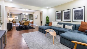 A modern living room with a sectional sofa, a dining area, and a kitchen in the background. - Spacious, Elegant Home w/ Cozy Backyard! (Troy)