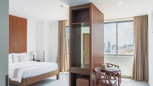 A hotel room with a bed, a desk, a chair, a mirror, and a large window with a city view. - The ONRA Hotel (Phnom Penh)