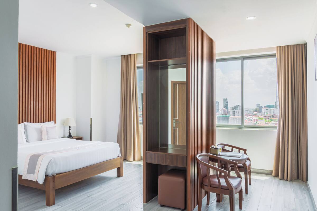 A hotel room with a bed, a desk, a chair, a mirror, and a large window with a city view.
