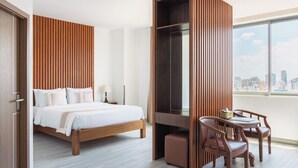A hotel room with a bed, a desk with chairs, a large window with a city view, and a wooden panel wall. - The ONRA Hotel (Phnom Penh)