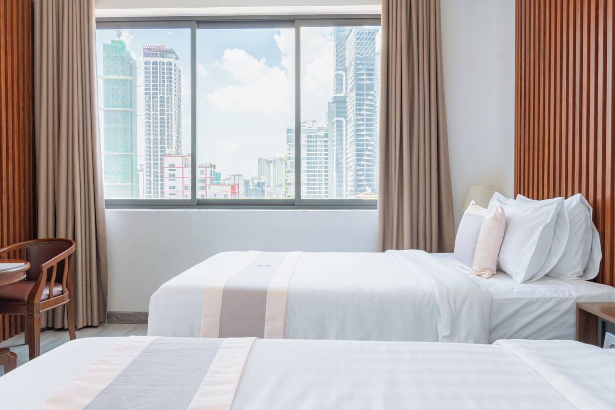 A hotel room with two beds, a view of the cityscape, a desk, and a chair.