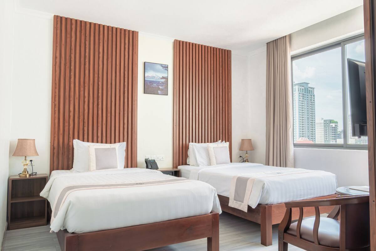 A hotel room with two beds, a desk, a chair, a window with a city view, and a mounted television.