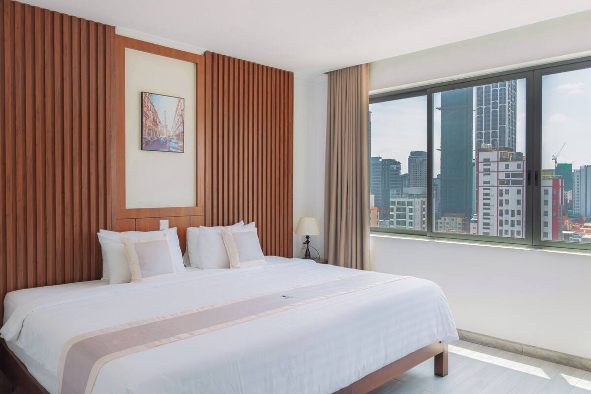 A hotel room with a large bed, a city view, a wooden panel wall, and a framed picture.