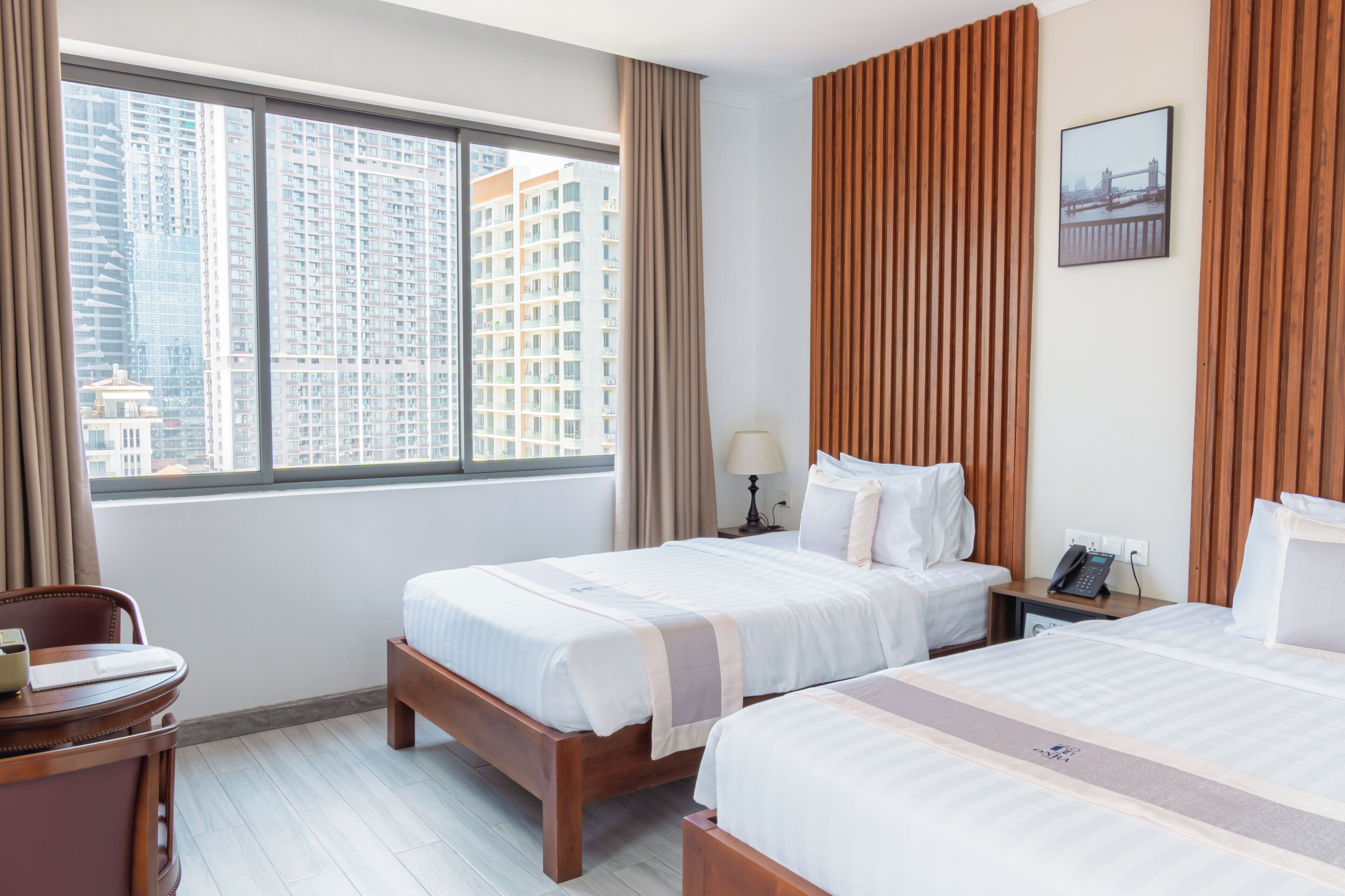 A hotel room with two beds, a desk, a chair, a window with a city view, and a wooden panel wall.