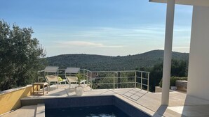 A swimming pool with a view of a hilly landscape, surrounded by a modern outdoor area with lounge chairs and a glass balustrade.