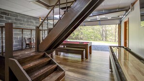 Una escalera de madera que lleva a una mesa de billar en una habitación con pared de piedra y vistas a árboles.