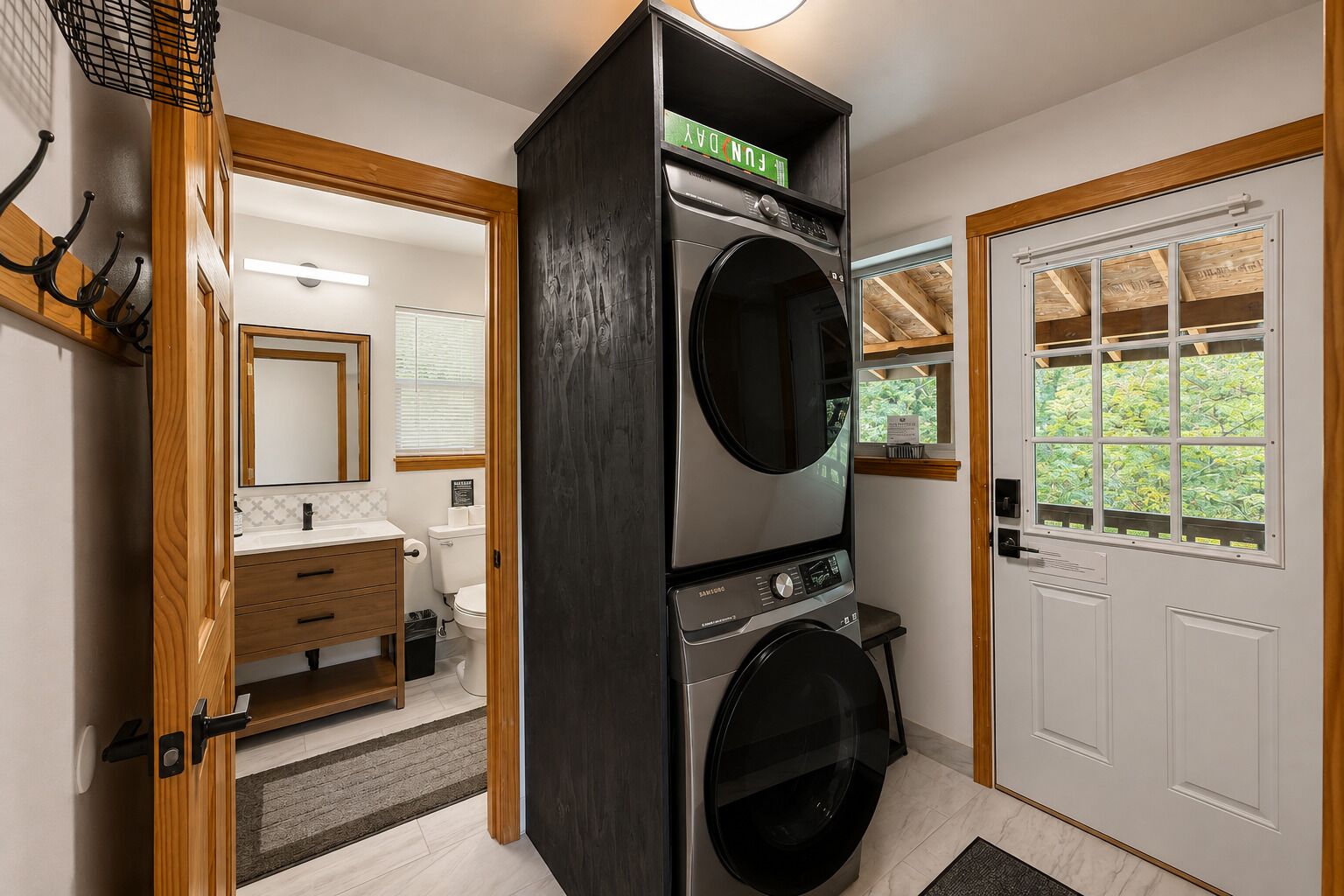 A laundry room with a front-loading washer and dryer, wooden cabinets, and a window with a view of greenery.
