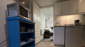 A narrow hallway with a blue shelving unit, a patterned rug, and a kitchenette area with cabinets and a refrigerator.