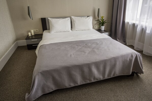 Un lit bien fait, recouvert d’une couvre-lit grise et agrémenté d’oreillers blancs, dans une chambre d’hôtel dotée d’un tapis gris, d’une table de chevet avec une lampe et d’une plante en pot.