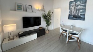 Un salon moderne avec un téléviseur à écran plat, des meubles blancs, une plante en pot et un tableau représentant un voilier accroché au mur.