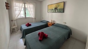 Une chambre avec deux lits, une fenêtre avec des rideaux légers, une petite table avec une lampe et un tableau au mur.