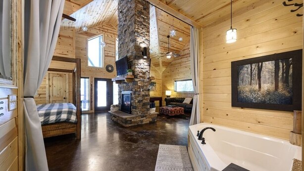 A log cabin interior with a stone fireplace, a large window, a cozy seating area, and a bathtub.