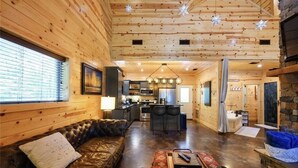 A cozy cabin interior with a leather sofa, a dining table, a kitchen area, and a stone fireplace.