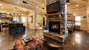 A spacious log cabin interior with a stone fireplace, a kitchen area, and a cozy seating area.