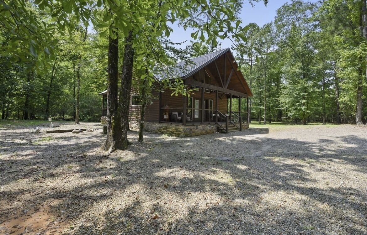 A rustic cabin with a porch, surrounded by trees and gravel ground.