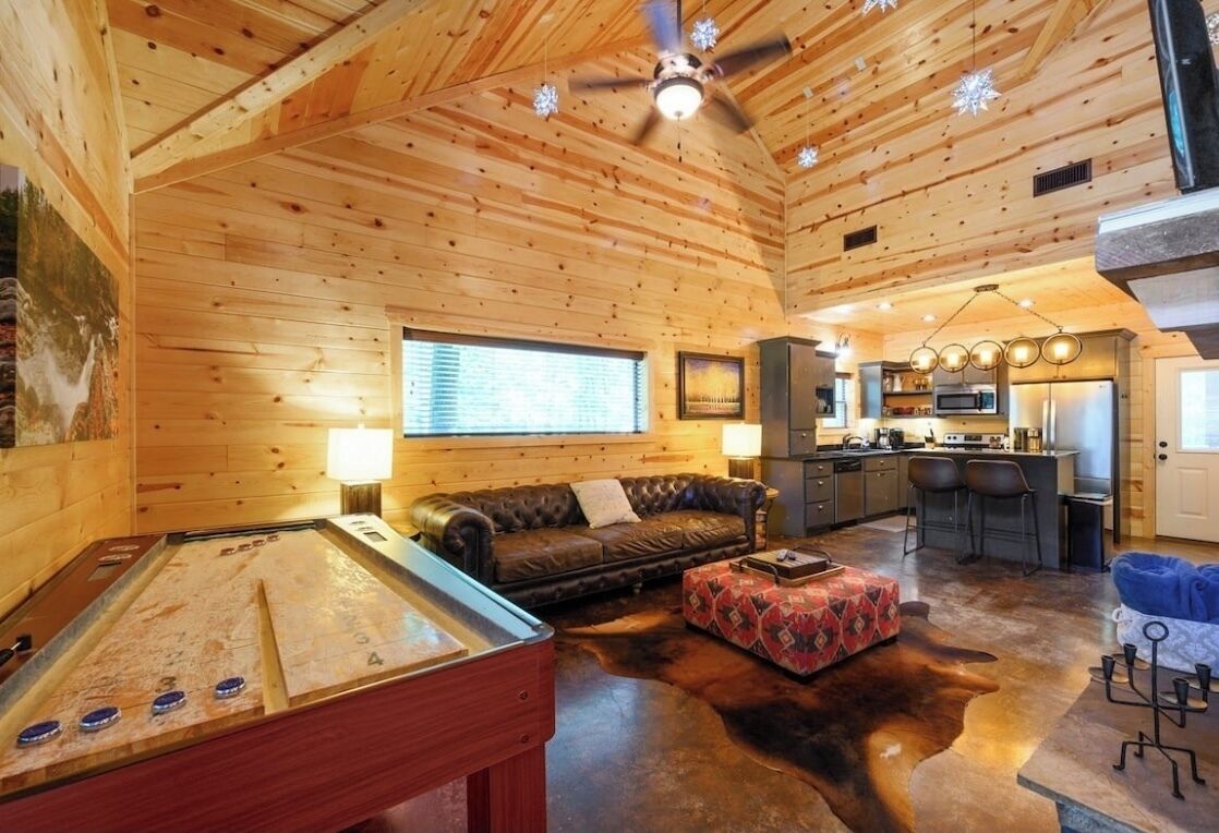 Acogedor interior de cabaña con techo de madera, mesa de billar, sofá y zona de cocina.