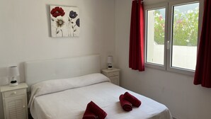 A bedroom with a bed, a nightstand, a lamp, a window with red curtains, and a floral artwork on the wall.