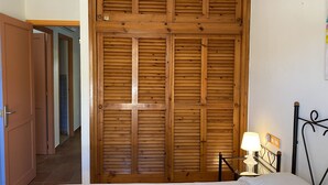 Une chambre avec une armoire en bois, un lit recouvert d’une courtepointe blanche, une table de chevet avec une lampe et une porte donnant sur un couloir.