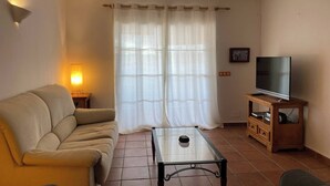 Un salon comprenant un canapé, une table basse en verre, un téléviseur posé sur un meuble en bois et une fenêtre avec des rideaux.