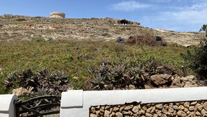 Une colline rocheuse surmontée d’une structure circulaire, un mur de pierre au premier plan et une végétation clairsemée.