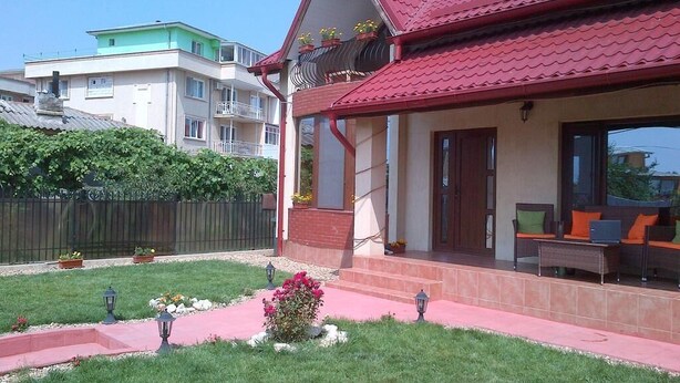 A two-story house with a red roof, a balcony with potted plants, and a covered porch with outdoor furniture.