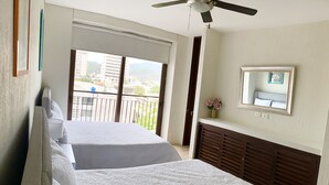 A hotel room with two beds, a ceiling fan, a large window with a view of buildings and mountains, a mirror, and a small vase with flowers. - Beautiful Executive Penthouse (Santa Marta)