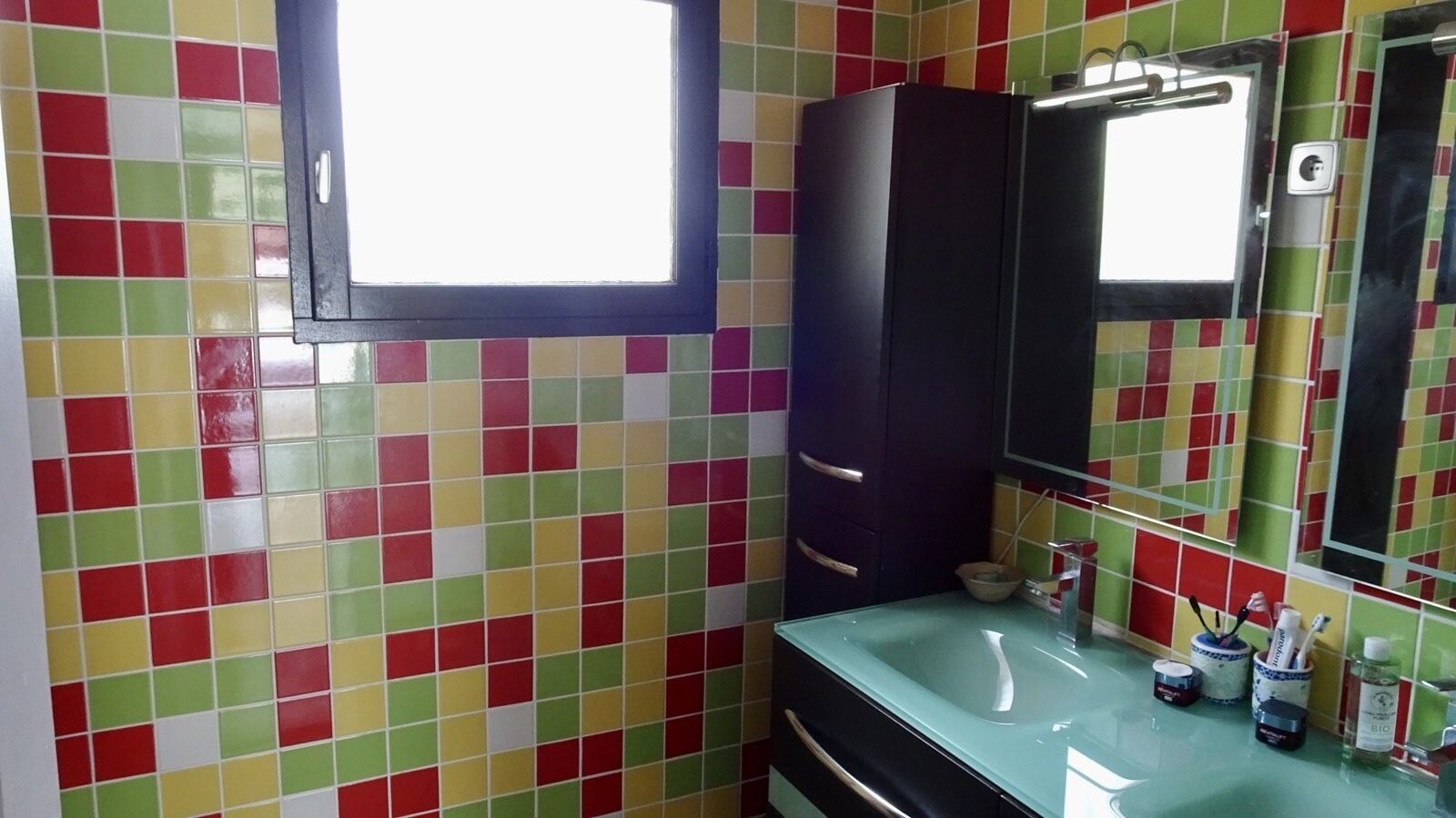Une salle de bain avec des carreaux de mosaïque aux couleurs vives, un meuble sombre, un lavabo avec un miroir et des articles de toilette posés sur le comptoir.