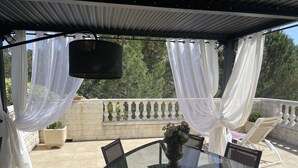 A covered outdoor patio with a glass table and chairs, white curtains, and a view of trees. - House with pool next to lake Saint-Cassien in the Esterel valleys. 20 min Cannes (Montauroux)
