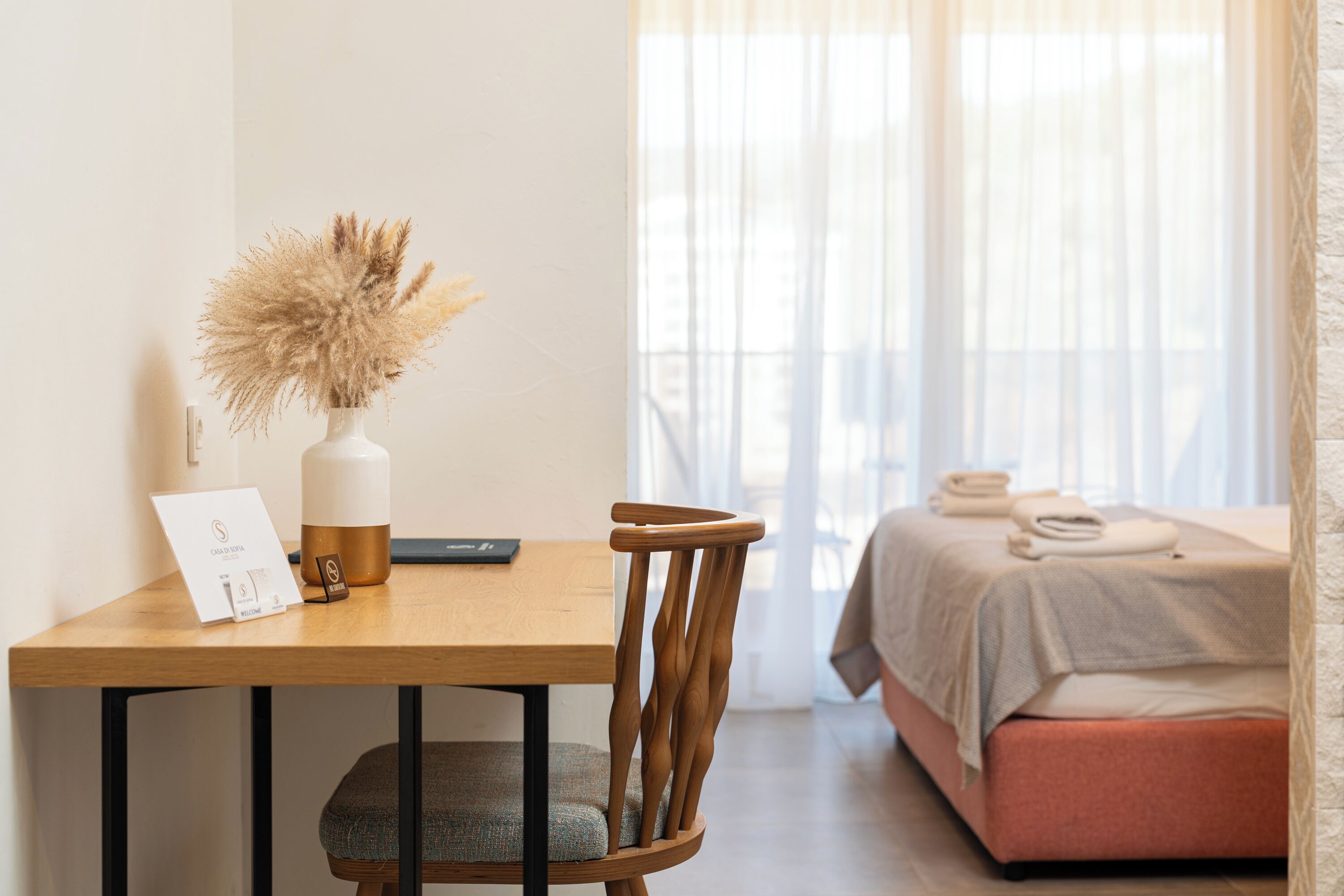 Une pièce avec un bureau en bois, un vase contenant des fleurs séchées, un lit avec un matelas rose et une fenêtre ornée de rideaux légers.