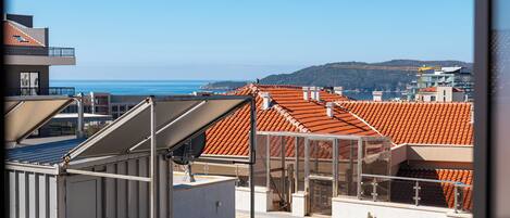 A rooftop terrace with a view of the sea, buildings with red-tiled roofs, and a construction crane in the distance.