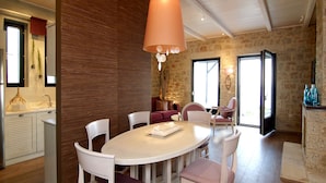 A dining area with a white table and chairs, a hanging light fixture, and a stone wall. - Enjoy the seaside living in a centraly located, pretty sea view house (Stoupa)