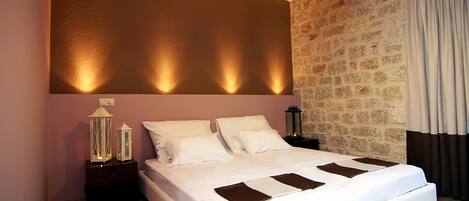 A neatly made bed with white linens and brown accent pillows, a wooden headboard, and a stone wall with a curtain on the right.