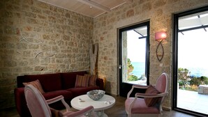 A cozy room with a stone wall, a red sofa, two pink chairs, a white table, and a view of the sea through a glass door. - Enjoy the seaside living in a centraly located, pretty sea view house (Stoupa)
