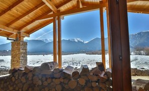 Una cabaña de madera con chimenea de piedra, vistas a montañas nevadas y un hoyo de fuego con leña.