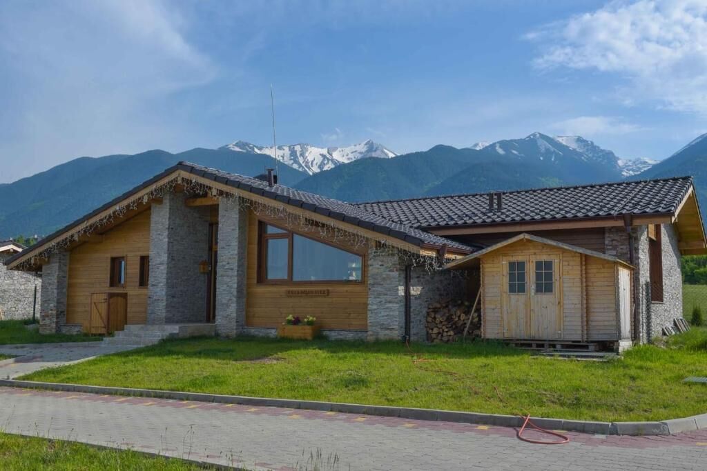 A stone house with a wooden exterior, a large window, and a mountain backdrop.