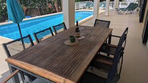 Een houten tafel met stoelen, een fles champagne en glazen op een terras met een zwembad en loungestoelen op de achtergrond.