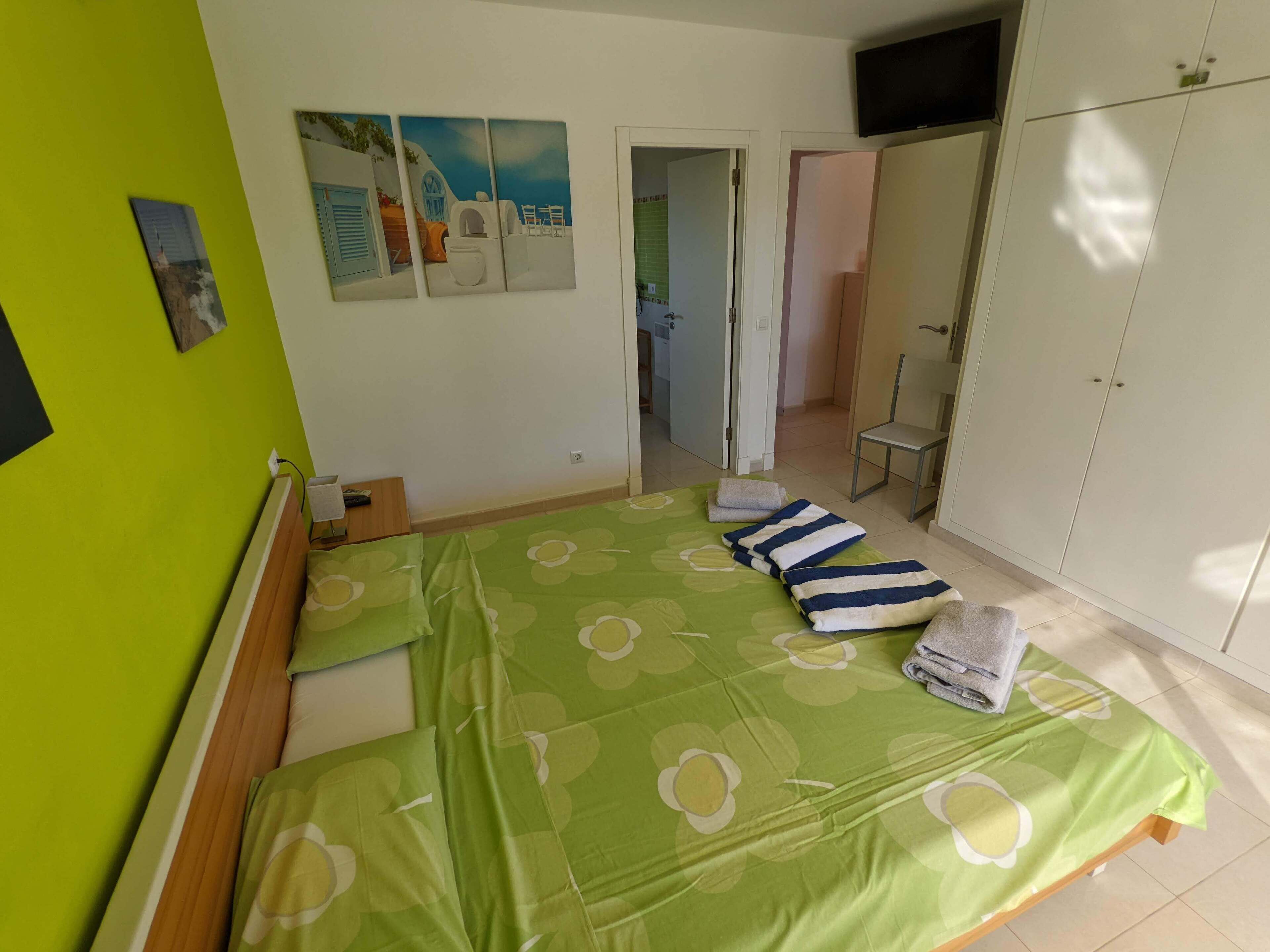 Quarto com cama, colcha floral verde, toalhas, uma cadeira, guarda-roupa e um quadro na parede.