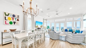 A spacious living area with a dining table, white chairs, a striped sofa, and a large window offering a view of the ocean.