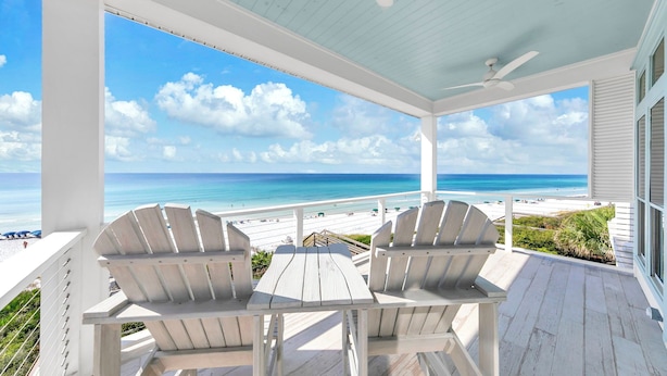 A covered outdoor seating area with two Adirondack chairs and a table, overlooking a beach with a clear blue sky and ocean.