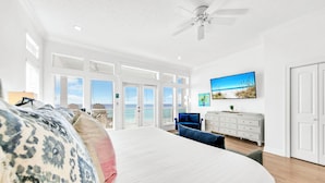 A spacious bedroom with a large bed, a ceiling fan, a dresser, and a painting of a beach scene.