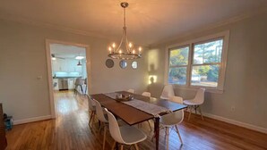 Un'ampia sala da pranzo con un tavolo in legno e sedie bianche, un lampadario e vista sulla cucina tramite un'apertura.