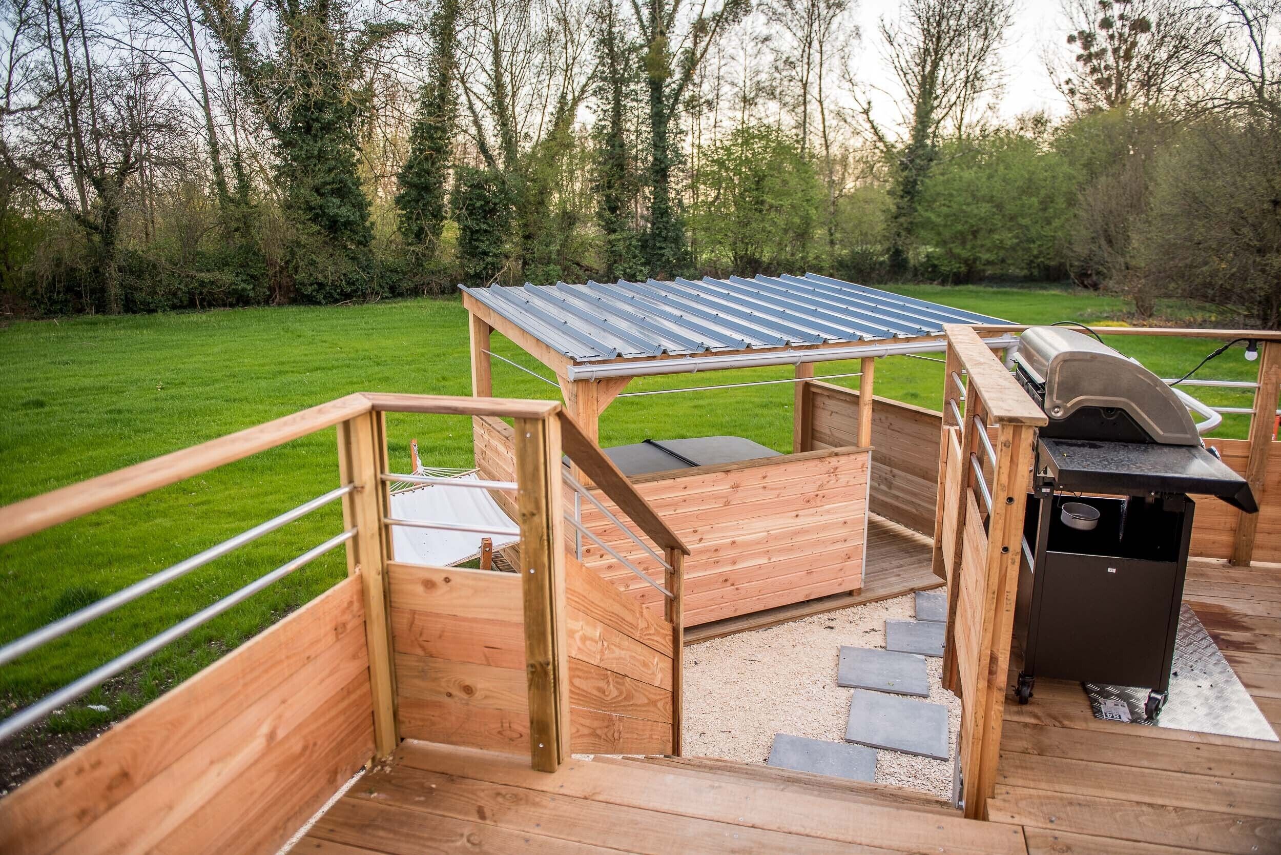 A wooden deck with a metal roof structure, a grill station, and a staircase leading to a raised platform.
