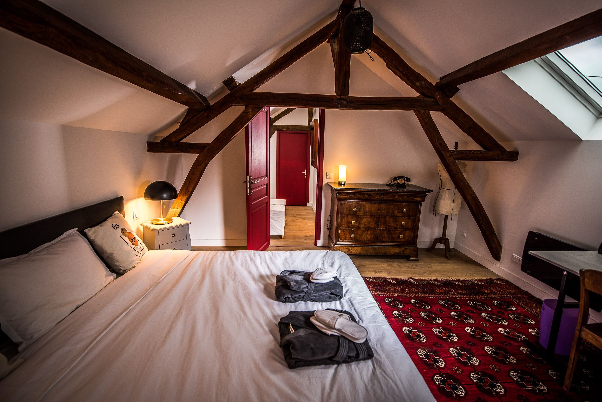 Une chambre avec un plafond en pente, un lit avec des oreillers, une table de chevet avec une lampe, une commode et une porte rouge en arrière-plan.